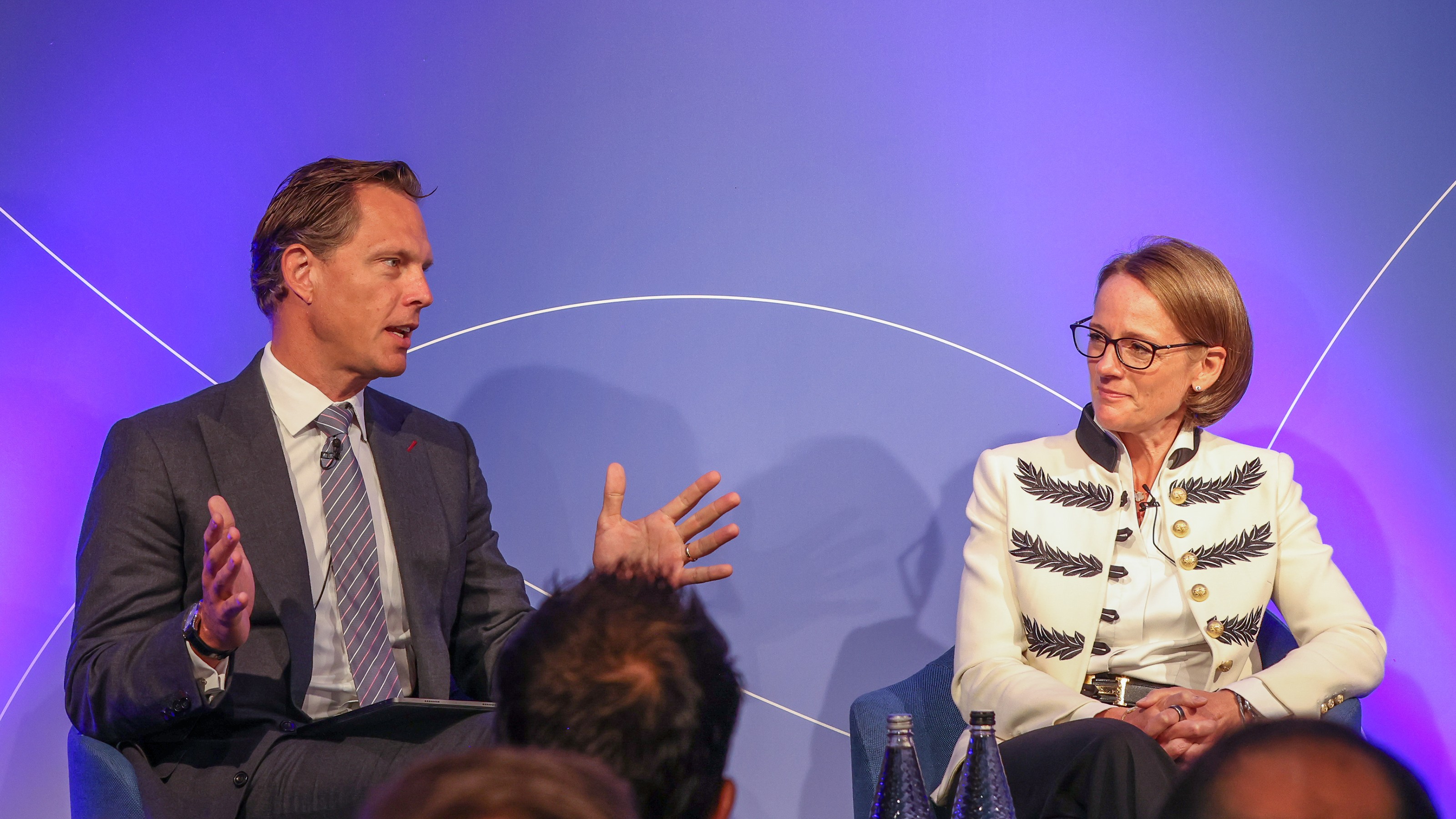 Two individuals seated on stage in conversation against a purple backdrop at a professional event.