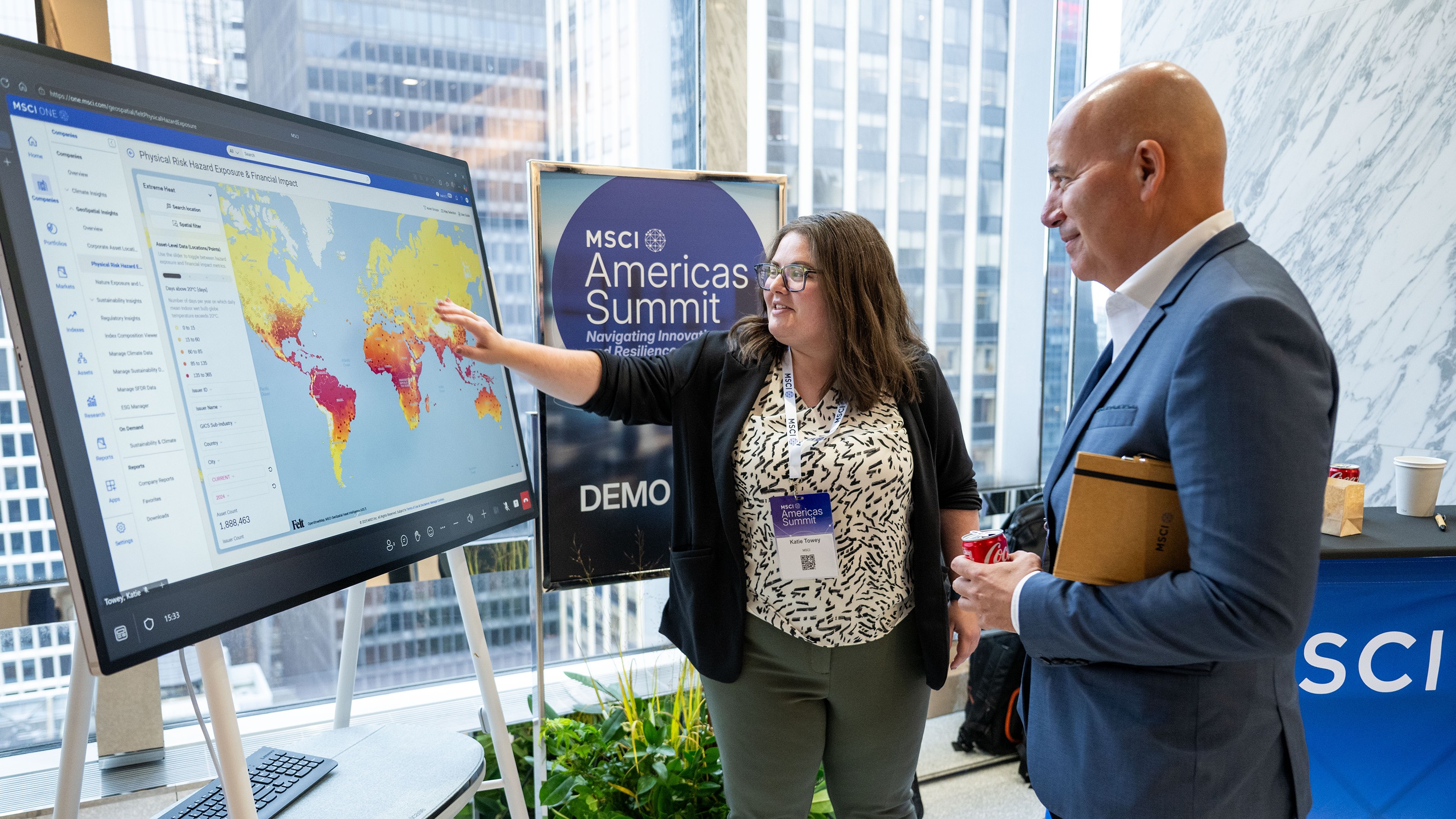 A woman points to a digital world map display while explaining data to a man at the MSCI Americas Summit.